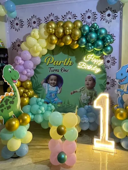 A cute dinosaur-themed first birthday party. This home setup includes a colorful balloon arch, a personalized backdrop with the baby's photos, and friendly dinosaur cutouts.