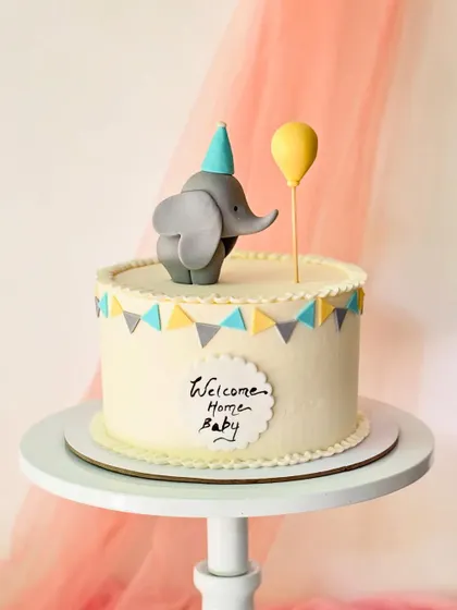 A "Welcome Home Baby" cake with a cute elephant topper. The simple design includes a yellow, grey, and blue bunting banner.