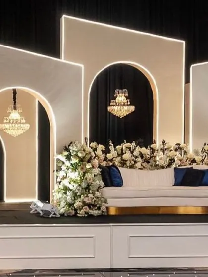A modern and sophisticated reception stage. The design features clean white geometric arches with built-in lighting, crystal chandeliers, and elegant white floral decor.