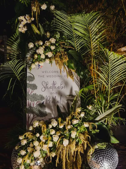 A chic welcome sign for a glamorous reception, surrounded by white roses, lush greenery, and sparkling disco balls.
