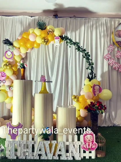 A naming ceremony setup with a yellow and pink balloon arch on a grass backdrop.