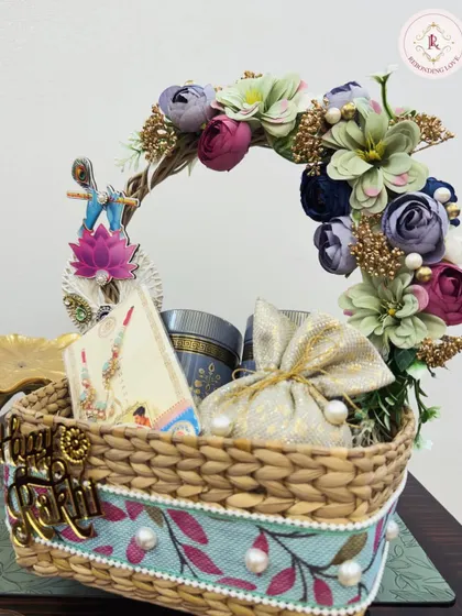 This elegant Rakhi basket features a beautiful floral handle and contains curated gifts like decorated mugs and potlis. The "Happy Rakhi" sign adds a final festive touch.