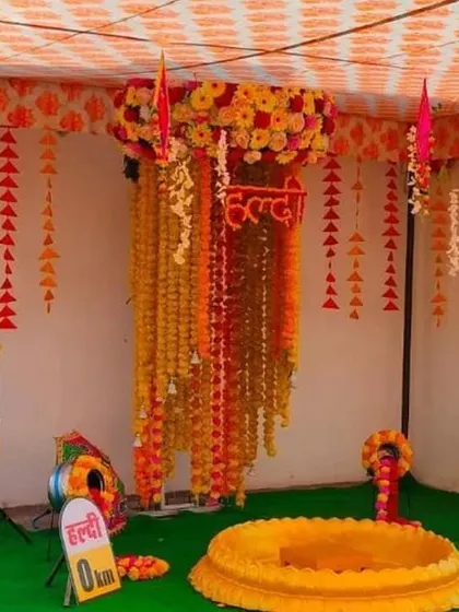 A creative Haldi setup with a canopy of hanging marigold garlands and fun "Haldi 0 km" milestone signs.