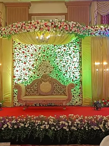 A traditional reception stage with a mix of floral walls and rich golden drapery. The layered look and classic furniture create a grand and festive atmosphere for a wedding celebration.