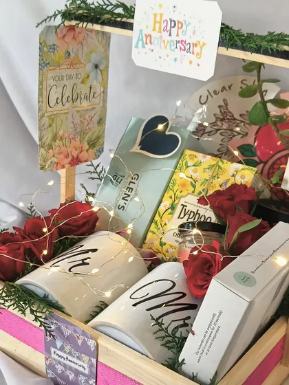 A perfect anniversary gift hamper to celebrate a milestone. This beautiful arrangement includes "Mr" and "Mrs" mugs, tea, snacks, and a personalized card, all lit up with fairy lights.