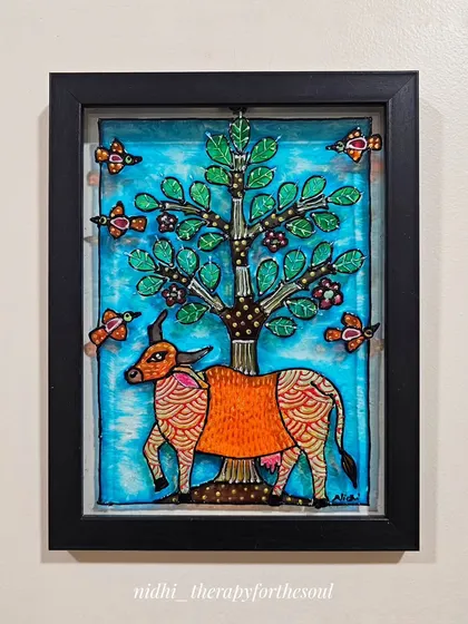 A framed Gond-style cow painting on glass. The cow stands under a tree with birds, showcasing a playful and colorful interpretation of the traditional art form.