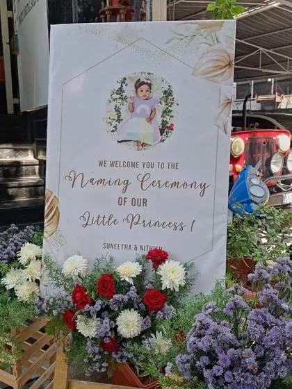A personalized welcome board for a little princess's naming ceremony. The board features a photo of the baby and is surrounded by fresh flowers.
