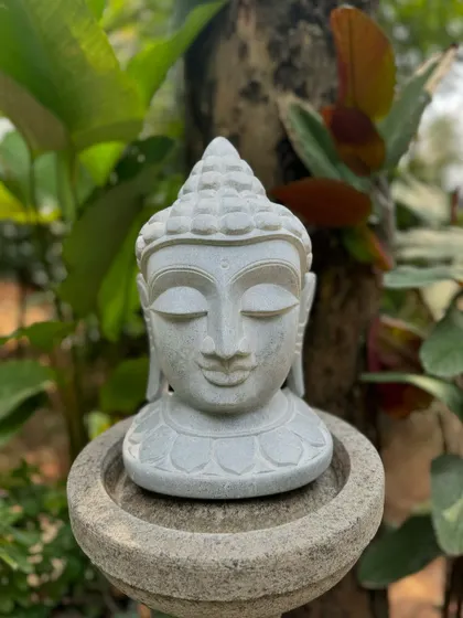 This stone Buddha bust, placed on a simple stone pedestal, invites reflection and harmony amidst the natural beauty of a garden.