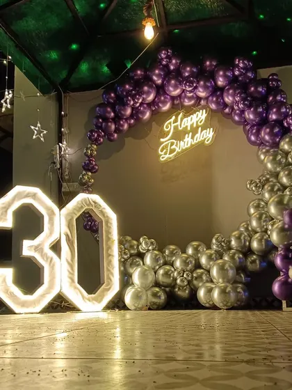 A dramatic 30th birthday on a terrace. The half-arch of metallic purple and silver balloons, combined with the large marquee numbers and neon sign, looks stunning against the dark sky.