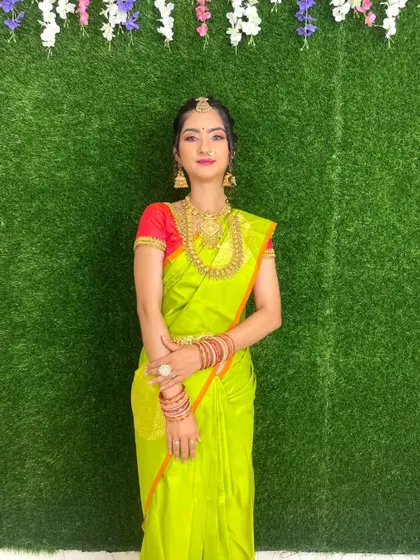 A classic pose for a South Indian bride against a simple green backdrop, allowing the vibrant saree, gold jewelry, and flawless makeup to stand out.