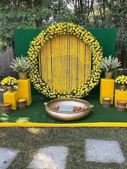 A clean and elegant outdoor Haldi setup with a circular marigold backdrop. The arrangement includes yellow pedestals with floral arrangements and a traditional brass 'urli'.