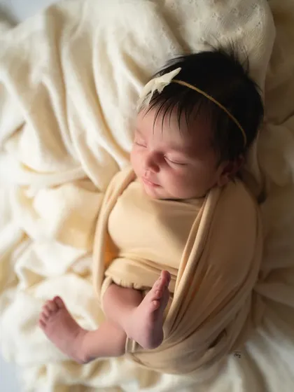 A simple and timeless portrait of a baby wrapped in a soft, butter-yellow swaddle, sleeping on a bed of cream-coloured fabric.