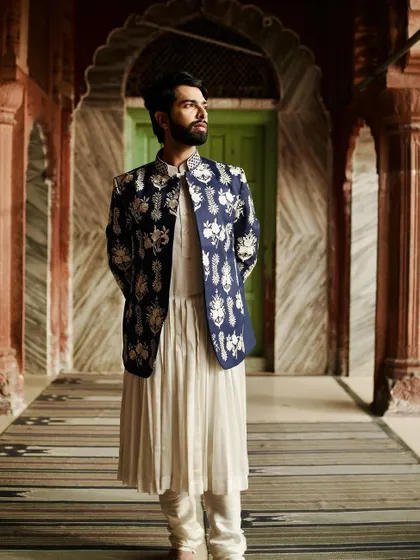 We love to reimagine royal aesthetics. This ensemble pairs a deep blue, richly embroidered jacket with a traditional pleated white kurta, creating a look of majestic elegance perfect for a wedding ceremony or grand reception.