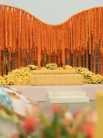A classic Haldi stage featuring a modern, undulating backdrop made entirely of orange marigold strings, with a comfortable yellow sofa for the couple.