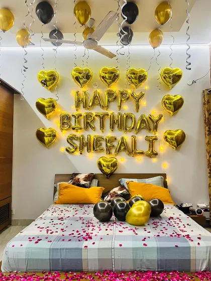 The bed is decorated with rose petals and balloons, completing the golden-themed birthday surprise.