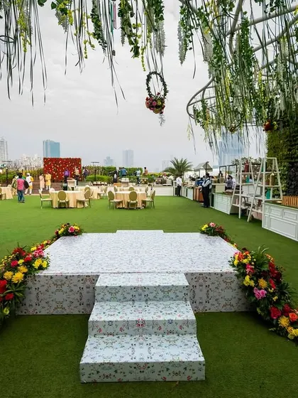 A daytime view of a rooftop venue, with a tiled stage accented by colorful red and yellow floral arrangements, set against the Mumbai skyline.