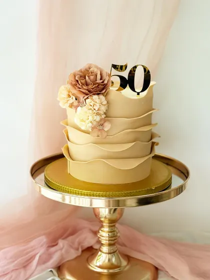 A sophisticated cake with a unique waved buttercream texture in a neutral beige tone. It's decorated with a "50" topper and silk flowers.