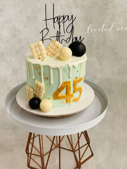 A sophisticated drip cake for a 45th birthday. The design uses a muted color palette with white chocolate drip, chocolate spheres, and a touch of gold leaf for a modern and stylish look.