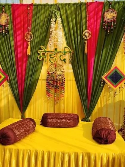 A Mehendi seating arrangement with a backdrop of green, pink, and yellow drapes, decorated with hanging dreamcatchers and tassels.