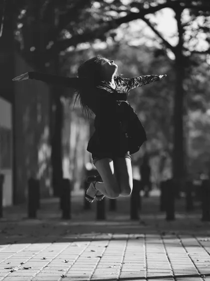 A joyful, dynamic black and white action shot of a woman jumping in the air with her arms outstretched. This image captures a moment of pure, uninhibited freedom on a tree-lined path.