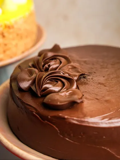 A close-up on the rich, glossy chocolate frosting of a cake. The focus here is entirely on the texture and the beautiful, handcrafted chocolate roses on top.