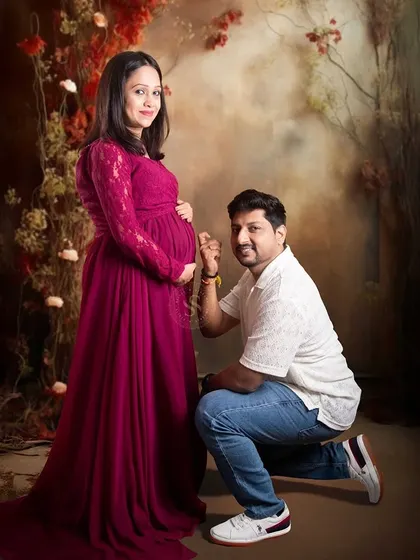A classic maternity pose with the partner listening to the baby bump, set against a floral-themed studio backdrop.