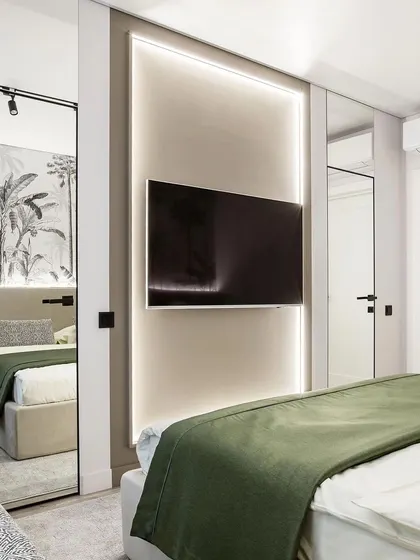 A bedroom with a TV mounted on a backlit panel, creating a soft glow. The room features a green bedspread and a mirrored wardrobe, making the space feel larger.