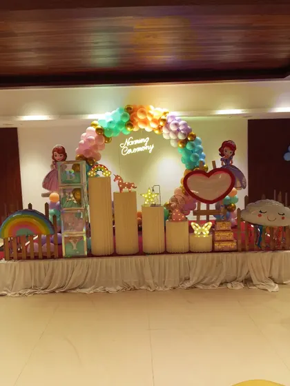 A rainbow princess theme for a Naming Ceremony. This setup features cutouts of Princess Sofia, a rainbow, a unicorn, and a multi-colored balloon arch over a 'Naming Ceremony' neon sign.