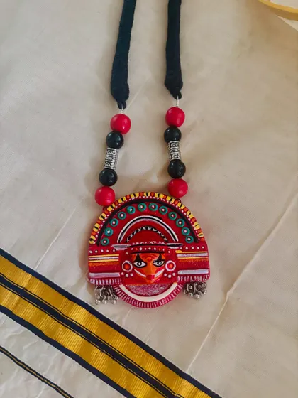 This terracotta pendant is inspired by the dramatic makeup of Theyyam performers. The bold red color and intricate details make it a vibrant statement piece, especially when paired with a traditional Kerala saree.