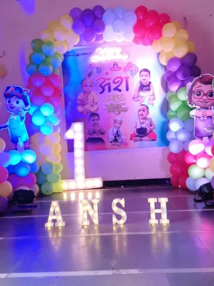 A complete Cocomelon stage setup for Ansh's first birthday. It features a custom backdrop with photos, character cutouts, and light-up letters for the name and number '1'.