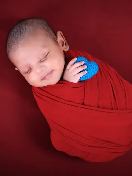 A sweet and simple shot of a smiling newborn holding a small blue heart. The focus is entirely on the baby's joyful expression and tiny hands.