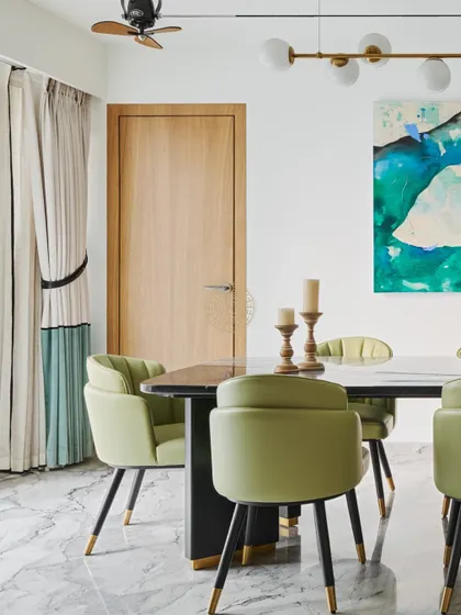 In this Mumbai duplex, the dining area is defined by soft sage green chairs and a marble-topped table. The abstract blue and green artwork on the wall adds a vibrant pop of color, tying in with the custom curtains.
