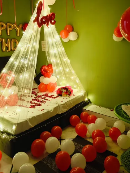 A birthday surprise with a fairy light canopy. The bed is decorated with rose petals and a balloon bouquet, with 'Happy Birthday' foil letters on the wall.
