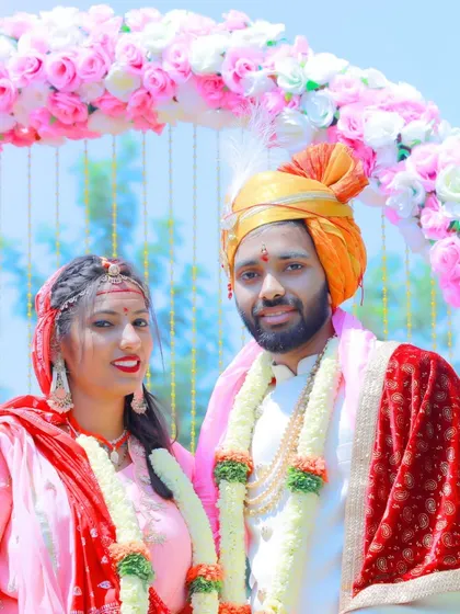 A traditional portrait of a couple in their wedding attire, standing under a beautiful pink floral arch.