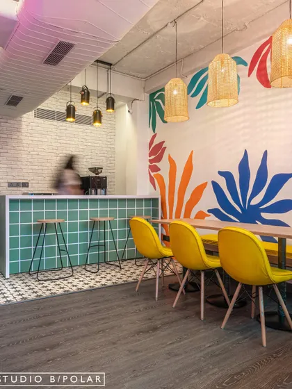 The cafeteria in the B-Hive workspace combines a fresh green tiled counter with bright yellow chairs and a vibrant, abstract wall mural. The space is designed to be bright and cheerful, providing a refreshing break from the workday.