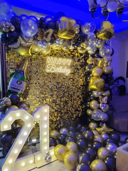 A stunning 21st birthday decoration with a gold shimmer wall. The setup is overflowing with black and gold balloons, a neon sign, and large marquee numbers.