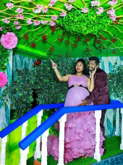 A duplicate of a playful maternity photo of a couple on our studio's garden bridge, pointing up at the whimsical floral ceiling.