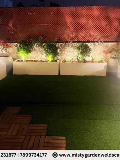 A beautiful night view of a transformed terrace garden. The artificial lawn is surrounded by illuminated planters and a PVC lattice screen for privacy, creating a magical evening ambiance.