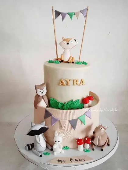 A stunning woodland animal themed cake for a first birthday. This two-tier cake features adorable, hand-crafted figures of a fox, raccoon, owl, and deer.