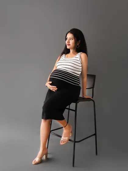A confident solo portrait of a mom-to-be in a modern striped outfit, seated on a high chair against a clean grey background.