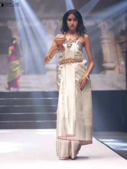 A striking pose under a spotlight at the NAVATAAR fashion event. The white saree with the gold belt creates a divine look, and the lighting adds a touch of theatrical drama.