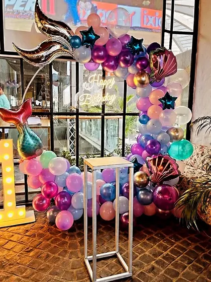 An enchanting mermaid-themed first birthday party. This decoration includes a balloon arch with mermaid tail foils, a 'Happy Birthday' neon sign, and a light-up number '1' against a large window backdrop.