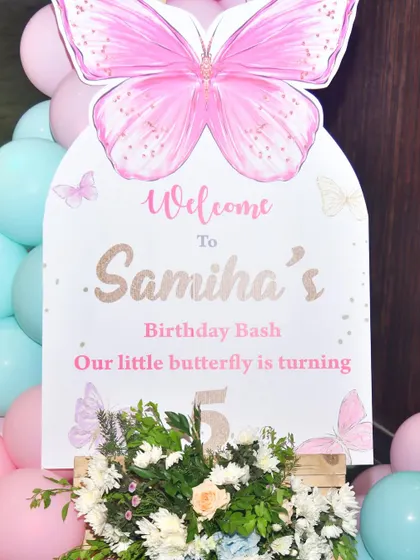 A close-up of a butterfly-themed welcome board for a 5th birthday bash. The sign is decorated with flowers and surrounded by pink and mint balloons.