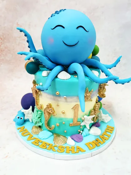 We explore the wonders of the ocean with this delightful blue octopus cake. A large, smiling octopus sits atop a sea-themed tier decorated with golden seahorses, shells, and other underwater treasures, perfect for a first birthday.