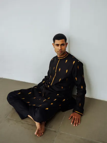 A minimalist shot for the Wabi Sabi menswear lookbook. The model is seated against a plain wall, drawing full attention to the black embroidered kurta.