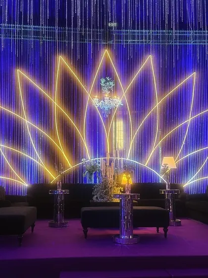 A stunning lounge area at the celestial-themed sangeet. A large neon lotus serves as the backdrop, complemented by elegant chandeliers and modern lamps, all under a canopy of deep blue and purple light.