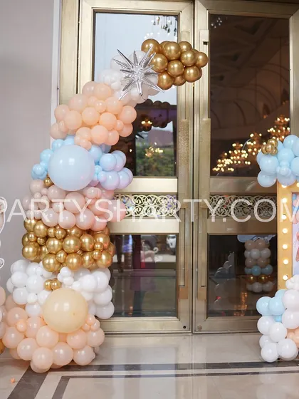 An elegant entrance decoration with a balloon arch in pastel peach, blue, and gold, complemented by a teddy bear cutout.
