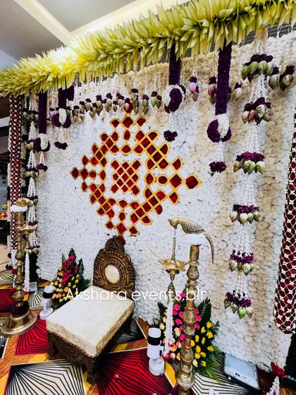 A beautiful backdrop for a Haldi or Sreemantham ceremony, made from a wall of white flowers with a geometric pattern in red. The decor is accented with hanging garlands and traditional lamps.