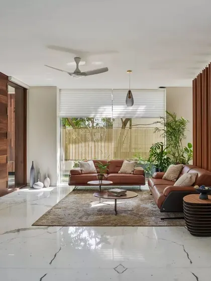 The formal living room at Kanasu, featuring clean lines, marble floors, and a warm leather sofa. The space is defined by a large wooden door and a slatted partition wall.