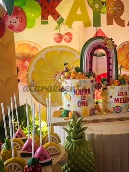 The dessert table is filled with on-theme treats, including adorable fruit-shaped cake pops that complement the main birthday cakes and the colorful backdrop.